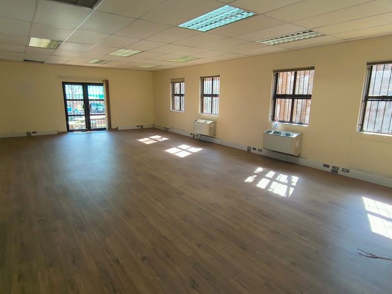 A spacious, empty office area featuring laminate flooring, recessed ceiling lights, and wall-mounted air conditioning units with security bars on the windows.