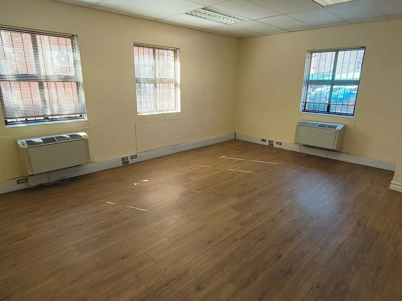 A bright, vacant office space featuring wood-look laminate flooring, suspended ceiling tiles, and dual wall-mounted air conditioning units.