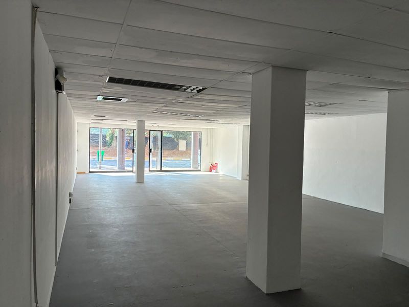 An empty open-plan retail space featuring a suspended ceiling, structural columns, concrete flooring, and large glass storefront doors providing natural light.