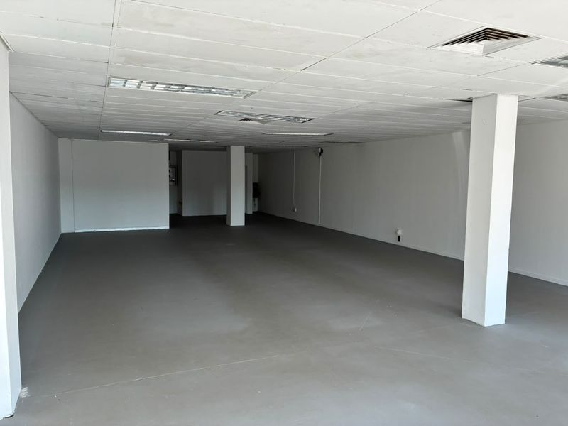 A spacious, vacant open-plan retail unit featuring a suspended ceiling, recessed lighting, air conditioning vents, and durable painted concrete flooring.