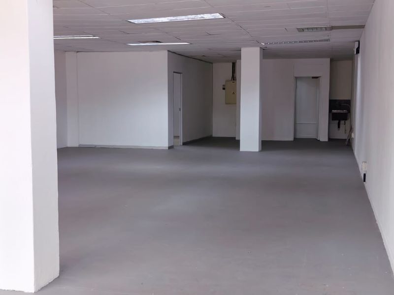 A spacious, open-plan retail unit featuring a neutral color palette, structural columns, suspended ceiling with recessed lighting, and a small utility sink area.