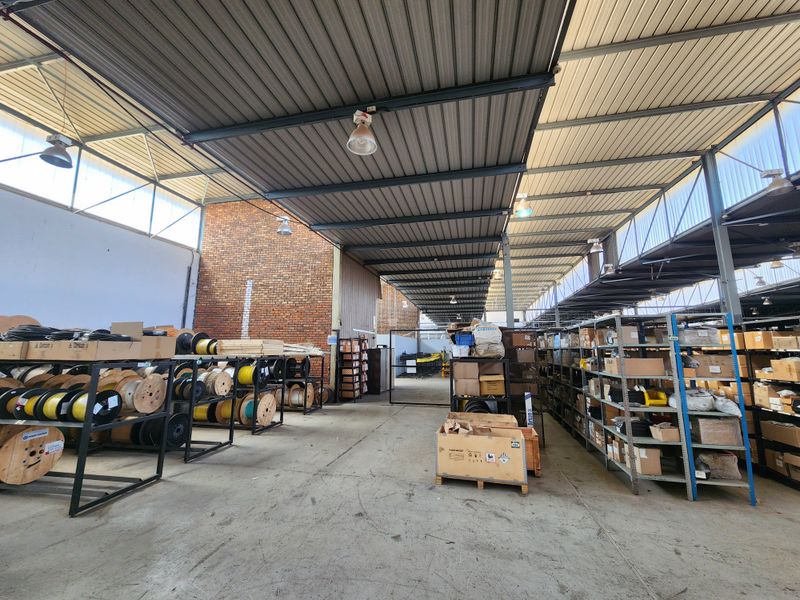 Spacious industrial warehouse area featuring high ceilings with exposed steel trusses, concrete flooring, and natural light through clerestory panels.