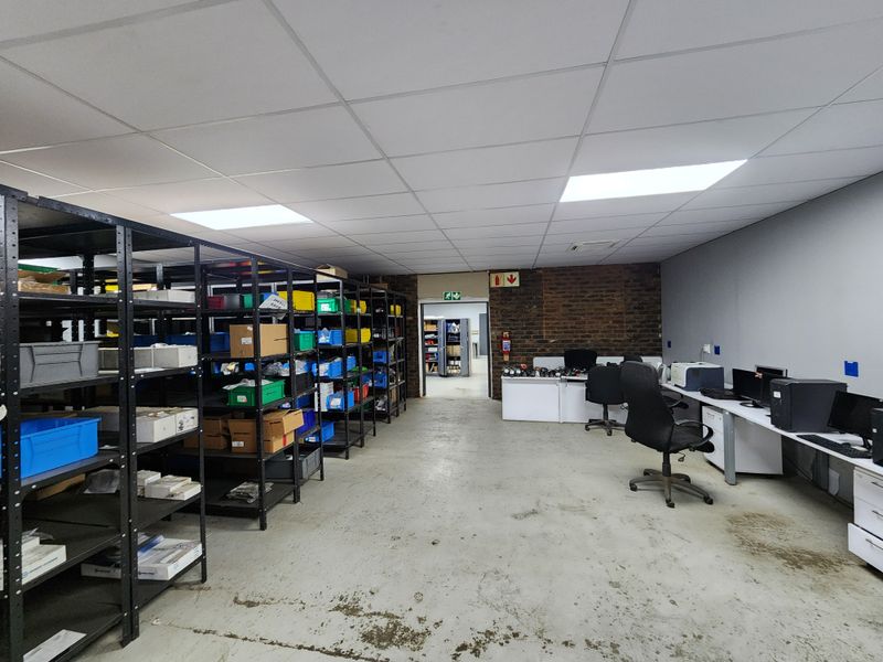 Industrial utility space featuring concrete flooring, suspended ceiling with recessed lighting, and an exposed brick feature wall, currently utilized for storage and office workstations.