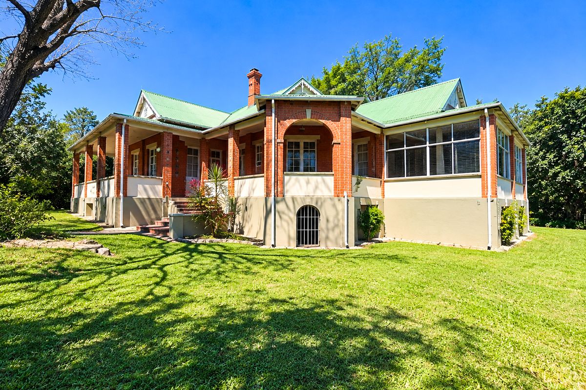 A well-maintained historic-style office building featuring red brick construction, a green metal roof, and a spacious covered veranda overlooking a large garden.