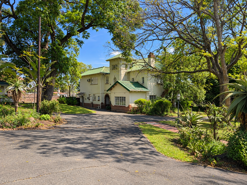 A well-maintained office building featuring a metal roof, brick foundation, and a spacious asphalt driveway surrounded by mature trees and landscaped gardens.