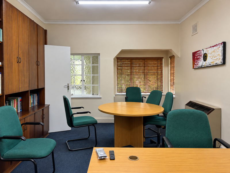 A functional office space featuring built-in wooden cabinetry, carpeted flooring, and natural light through windows with fitted wooden blinds.