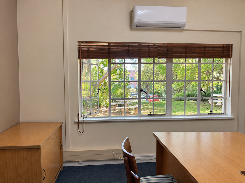 A bright office space featuring a split-system air conditioner, large multi-pane windows with timber blinds, and carpeted flooring overlooking a garden.