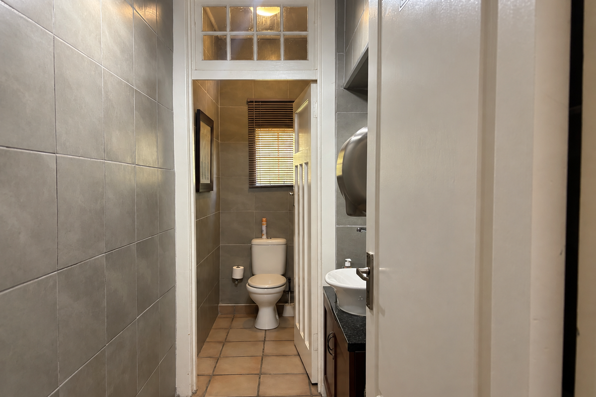 A functional bathroom featuring grey ceramic wall tiles, tiled flooring, a vanity with a vessel sink, and a separate toilet area with a transom window.