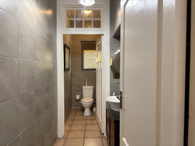 A functional bathroom featuring grey ceramic wall tiles, tiled flooring, a vanity with a vessel sink, and a separate toilet area with a transom window.