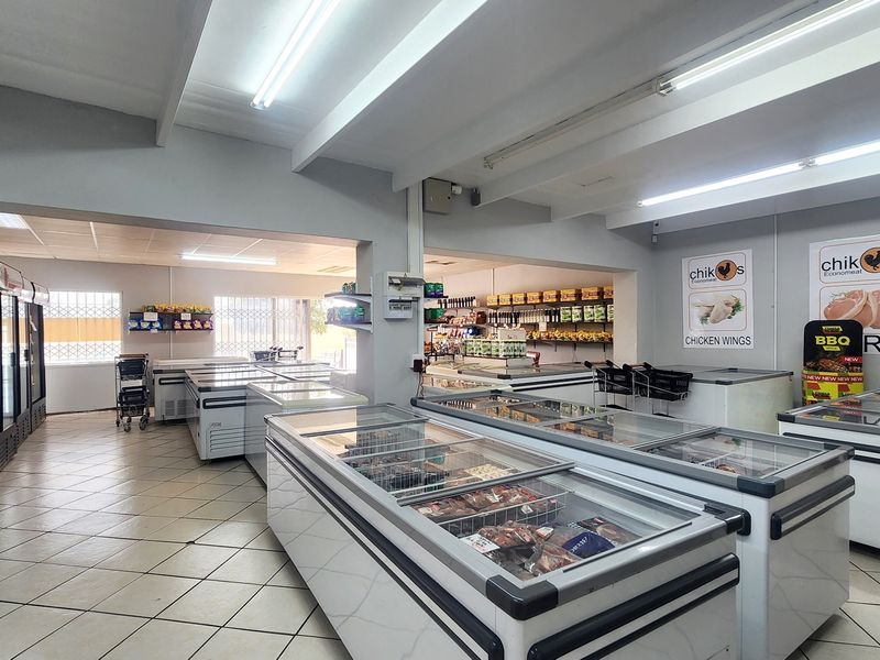 Spacious retail interior featuring tiled flooring, exposed ceiling beams, fluorescent lighting, and extensive built-in refrigeration units for commercial food display.