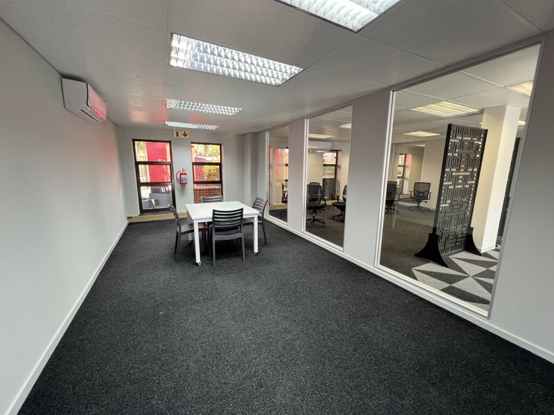 A modern office space featuring glass partitions, recessed lighting, wall-mounted air conditioning, and dark carpeting, providing a professional and functional work environment.