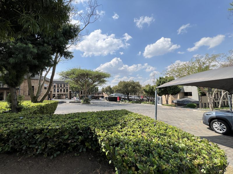 A well-maintained office park exterior featuring a paved driveway, mature landscaping, and covered parking bays under shade cloth structures.