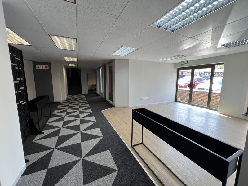 A bright, modern office space featuring a mix of patterned carpet and wood-look flooring, recessed ceiling lights, and glass doors leading to the exterior.