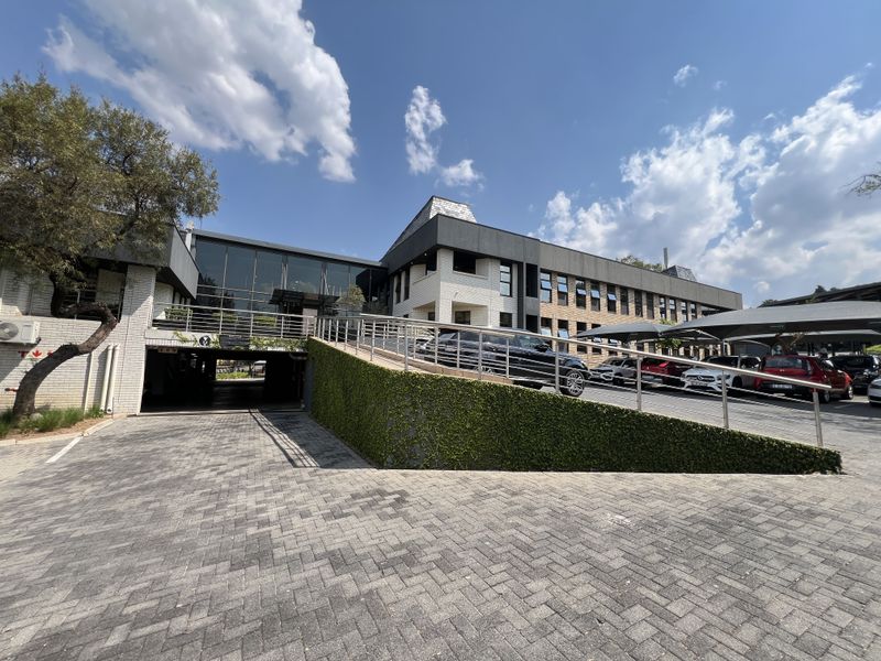 Front exterior view of a modern office building featuring a paved driveway, covered parking area, and a combination of brick and glass architectural elements.