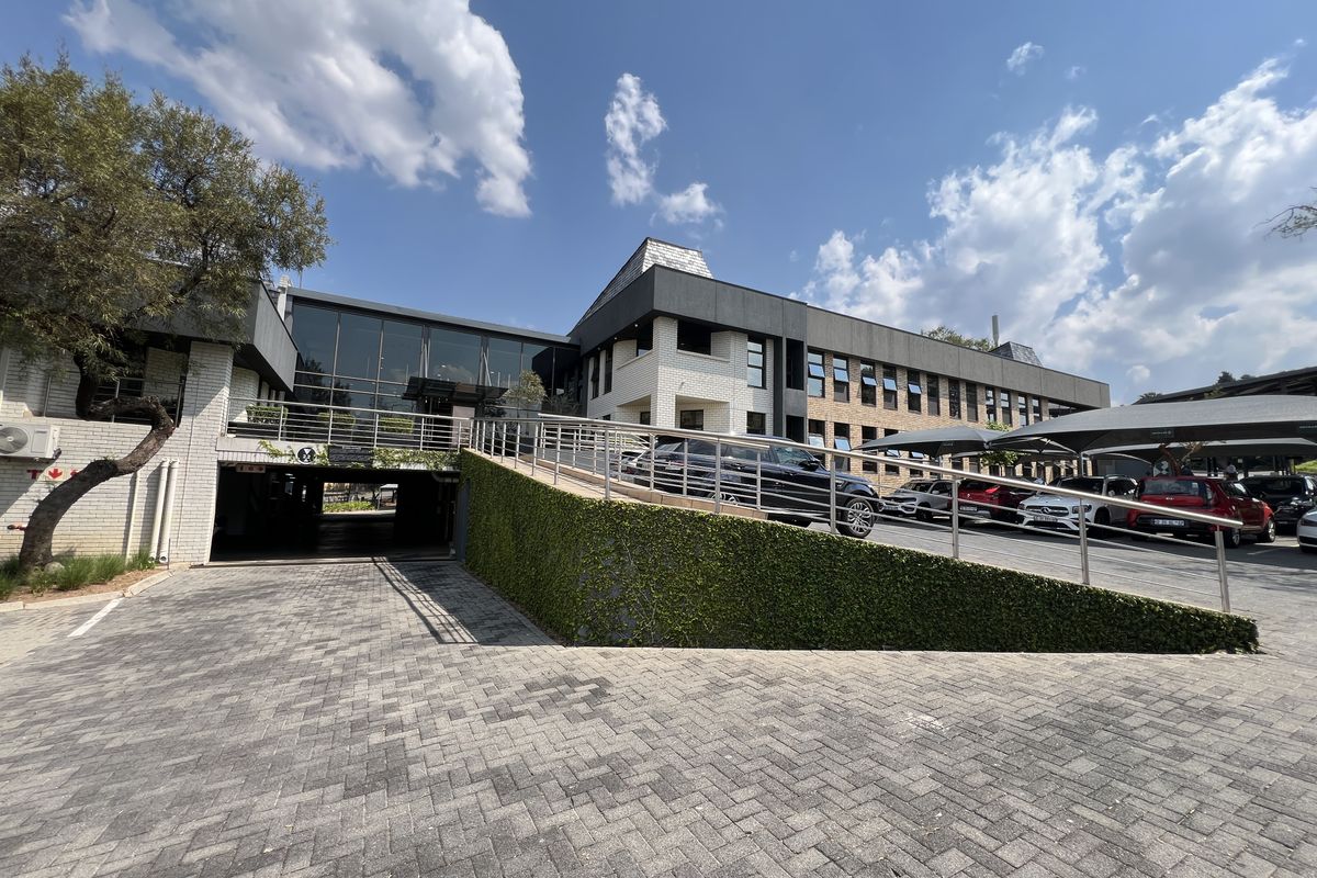 Front exterior view of a modern office building featuring a paved driveway, covered parking area, and a combination of brick and glass architectural elements.