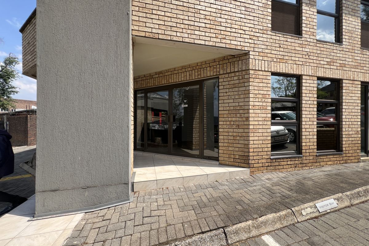 Front entrance of a modern office building featuring a face brick facade, aluminium glass doors, and a paved visitor parking area.