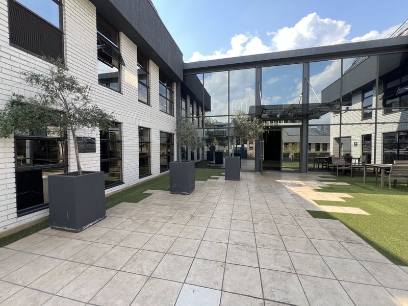 A modern commercial courtyard featuring a tiled patio, artificial turf, and extensive glass curtain walling connecting the building wings.
