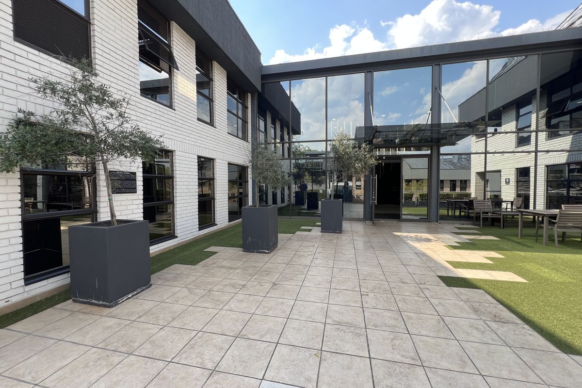 A modern commercial courtyard featuring a tiled patio, artificial turf, and extensive glass curtain walling connecting the building wings.
