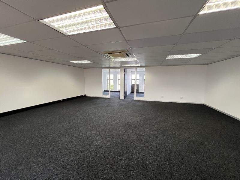 A spacious, empty commercial office area featuring a suspended ceiling with recessed lighting, dark carpeting, and glass-partitioned doorways leading to adjacent rooms.