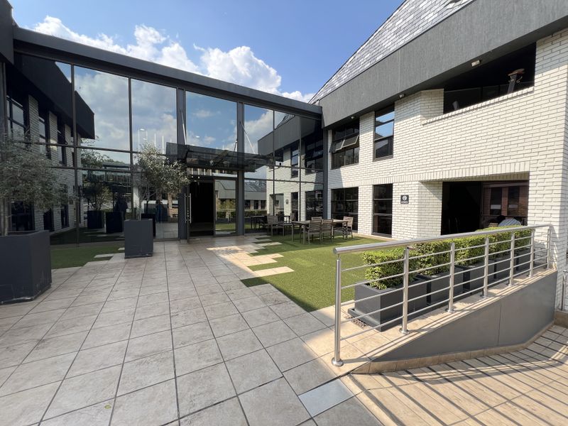 A modern commercial courtyard featuring a glass curtain wall, tiled patio area, artificial turf, and stainless steel railings, providing a professional outdoor breakout space.