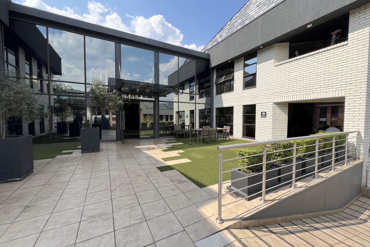 A modern commercial courtyard featuring a glass curtain wall, tiled patio area, artificial turf, and stainless steel railings, providing a professional outdoor breakout space.