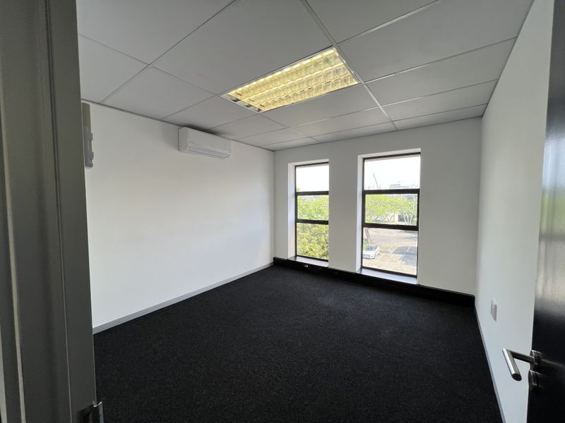 A clean, empty office space featuring dark carpeting, a suspended ceiling with recessed lighting, and a wall-mounted air conditioning unit.
