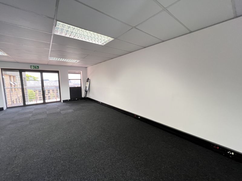 A bright, vacant commercial office space featuring a suspended ceiling, recessed lighting, carpet tiles, and sliding glass doors leading to a balcony.