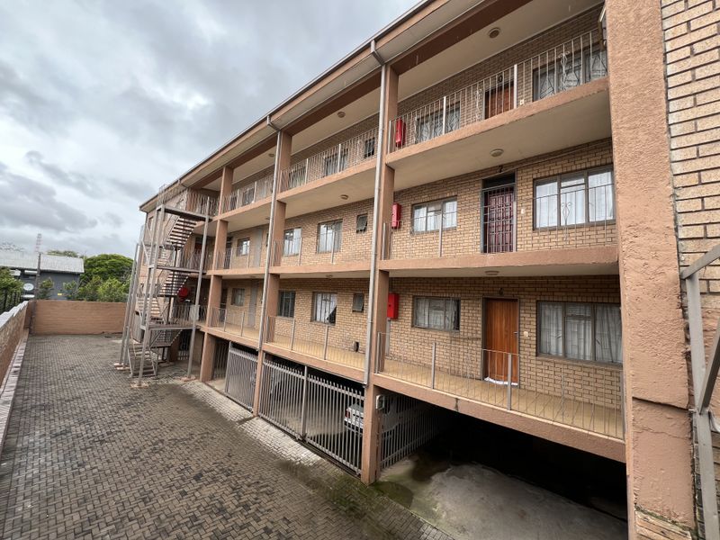 Exterior view of a multi-story apartment building featuring brick facade, private balconies, metal fire escape, and ground-level parking area.