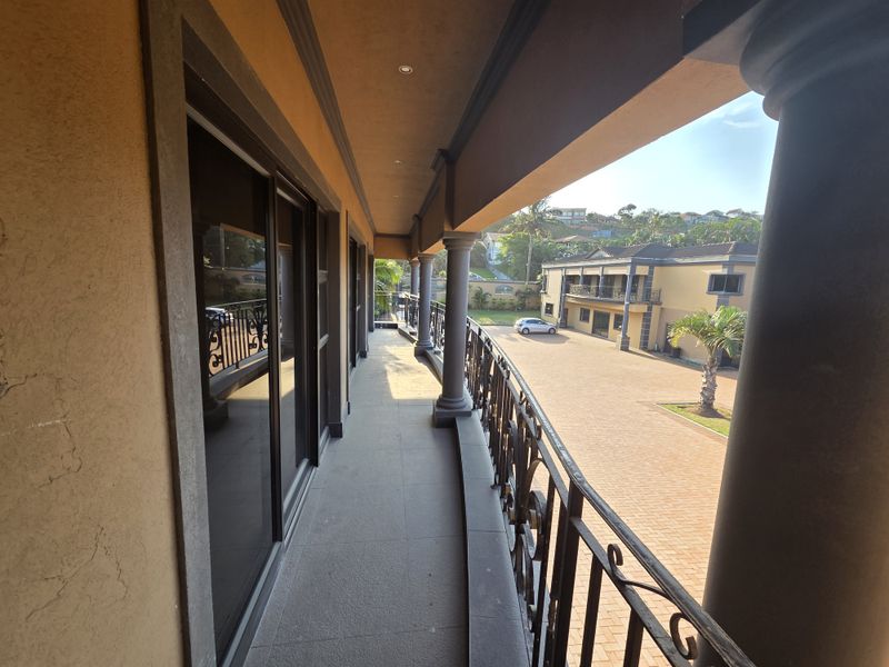 A spacious covered balcony featuring tiled flooring, wrought iron railings, and structural columns, offering an elevated view of the paved courtyard and surrounding property.