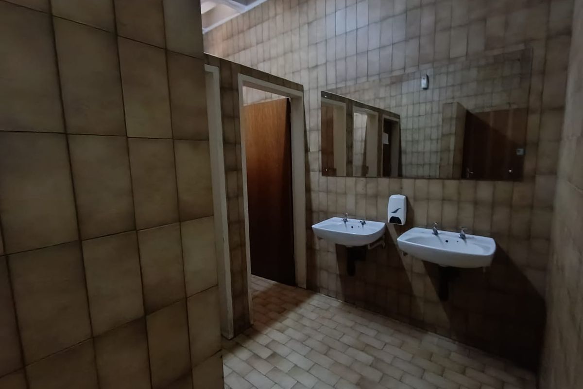 A functional commercial restroom featuring dual wall-mounted ceramic basins, tiled walls and flooring, and private cubicle access.