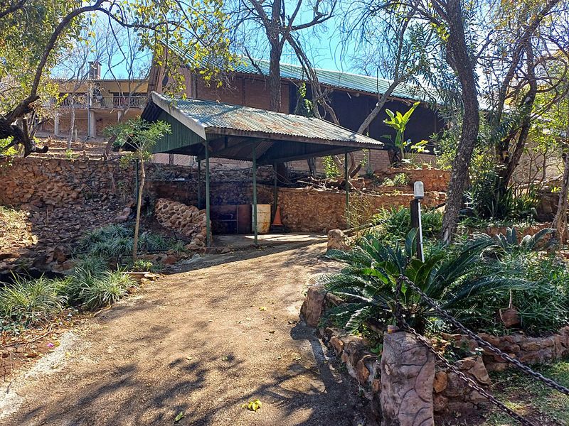 An outdoor covered patio area featuring a corrugated metal roof, stone retaining walls, and a concrete pathway set within a natural garden landscape.