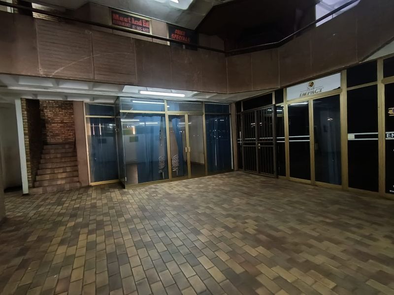 Interior view of a retail arcade or shopping center atrium featuring paved flooring, glass storefronts, and a mezzanine level with signage.