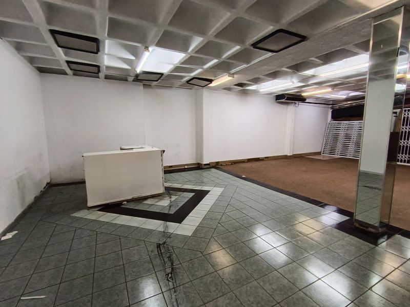 Open-plan retail space featuring a coffered ceiling, tiled flooring with decorative inlay, mirrored pillar, and fluorescent lighting.