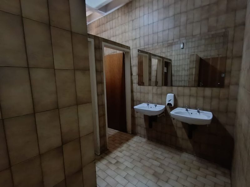 A functional commercial restroom featuring dual wall-mounted ceramic basins, tiled walls and flooring, and private cubicle access.