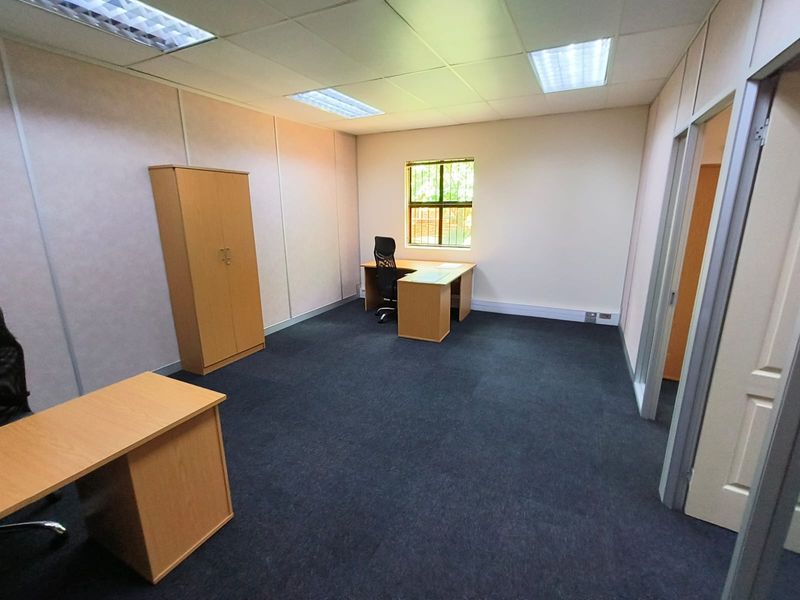 A functional office space featuring carpeted flooring, a suspended ceiling with recessed lighting, and a window equipped with security bars.