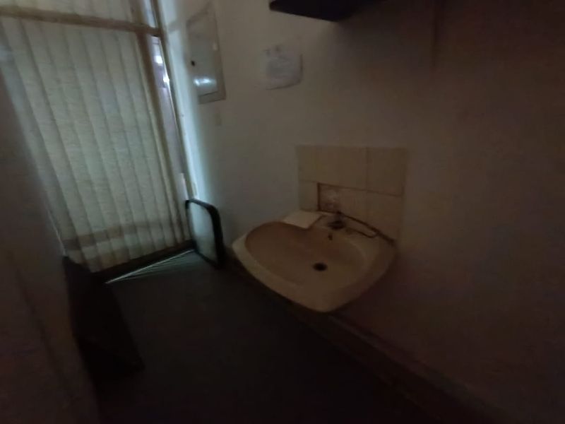 A small utility space featuring a wall-mounted sink with missing backsplash tiles and vertical blinds covering the window.