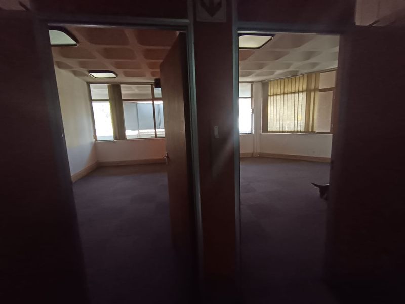 Two adjacent vacant office spaces featuring carpeted flooring, large windows with vertical blinds, and recessed ceiling lighting.
