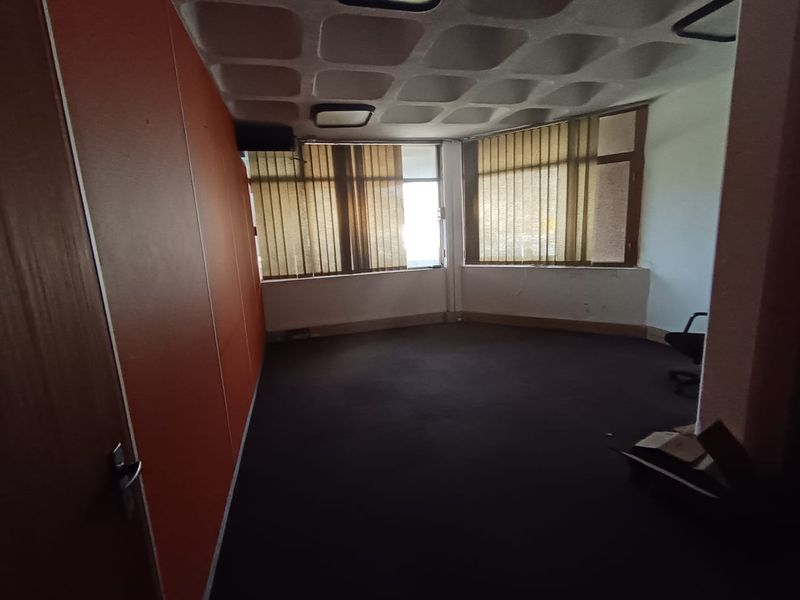A vacant office space featuring a distinctive waffle-patterned ceiling, large windows with vertical blinds, and dark carpeting, offering a blank canvas for professional use.