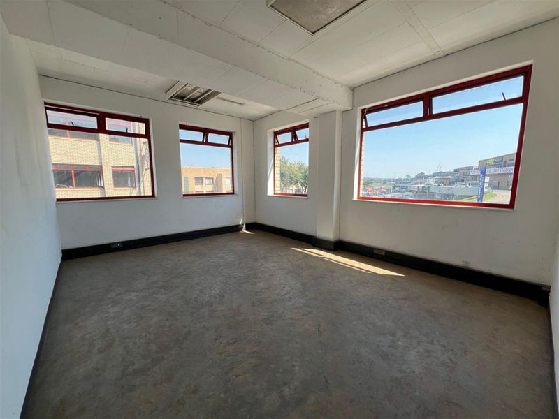 An empty, industrial-style office space featuring large windows with red frames, exposed concrete flooring, and a suspended ceiling.