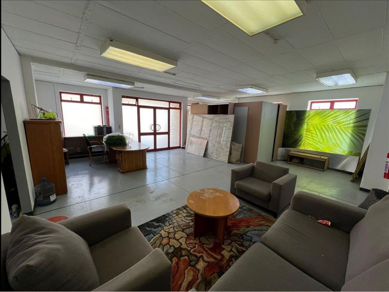Spacious industrial office area featuring concrete flooring, suspended ceiling with fluorescent lighting, and large aluminium-framed windows providing natural light.