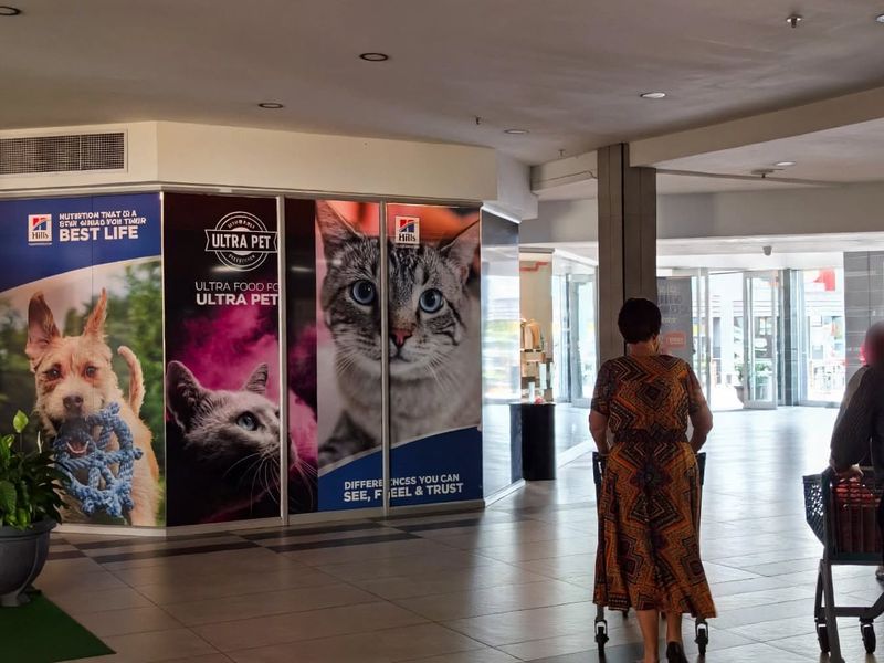 Interior view of a retail mall corridor featuring tiled flooring, recessed lighting, and a large glass storefront display with promotional branding.