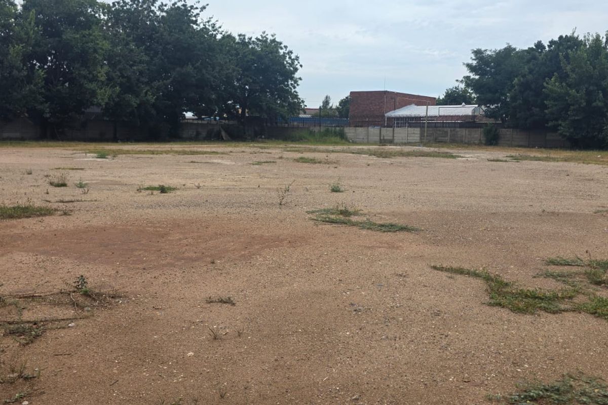 A wide, vacant commercial land plot featuring a level, unpaved surface enclosed by perimeter boundary walls and mature trees.