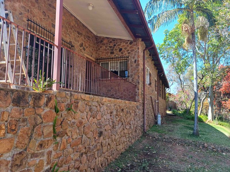 A side view of a stone-clad farmhouse featuring a covered patio area with metal railings and established garden surroundings.
