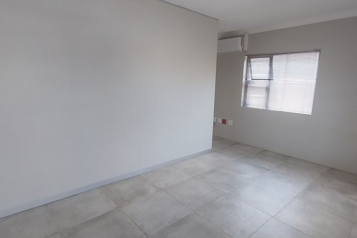 A clean, empty office space featuring neutral tiled flooring, an installed air conditioning unit, and window blinds, providing a versatile professional environment.