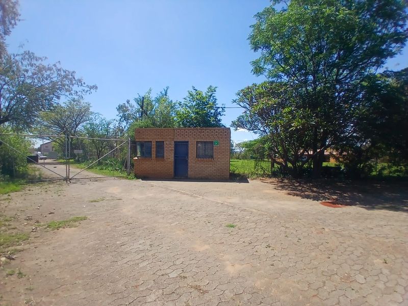 Industrial property entrance featuring a brick security guard house, metal gate, and paved driveway access.