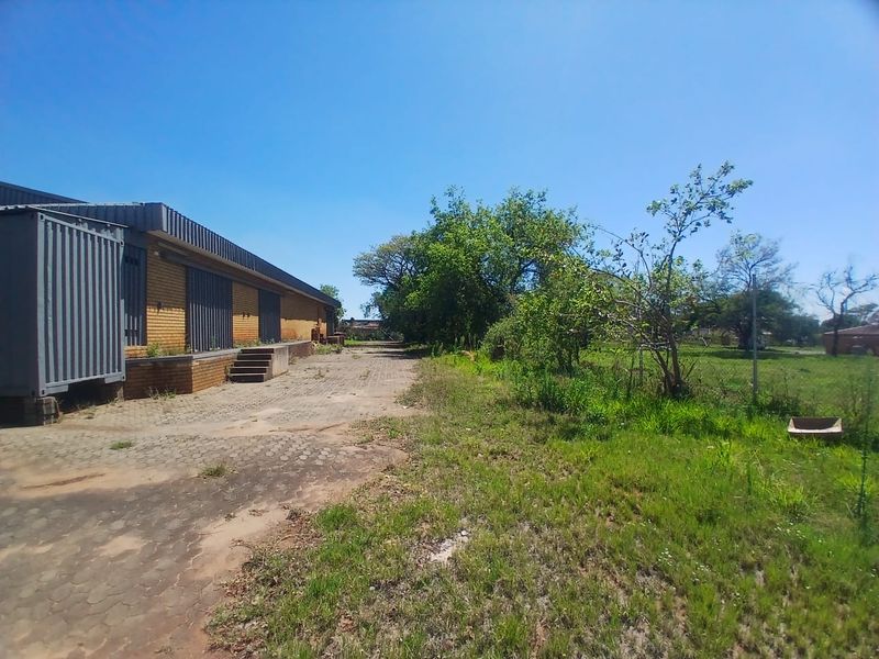 Rear exterior view of an industrial property featuring a brick facade, loading dock area, and paved yard space with surrounding vegetation.