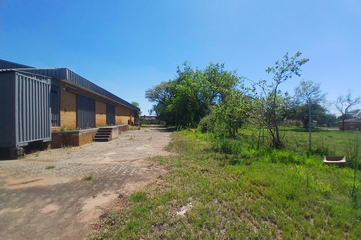 Rear exterior view of an industrial property featuring a brick facade, loading dock area, and paved yard space with surrounding vegetation.