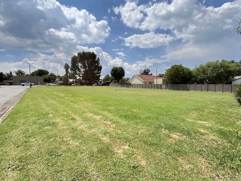 A wide-angle view of a vacant commercial land plot featuring level grass terrain, perimeter fencing, and direct street access in a developed area.