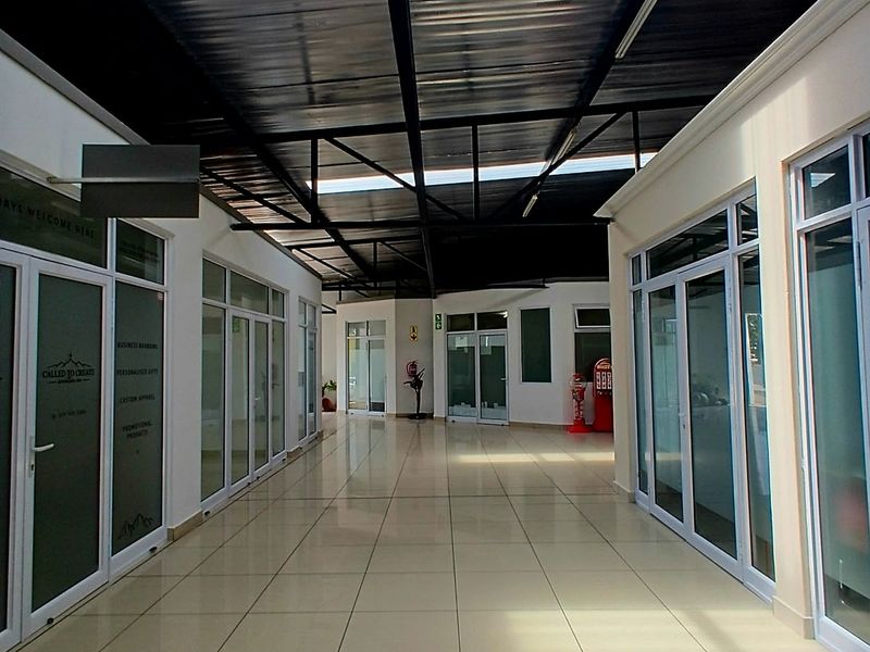 A bright, modern commercial hallway featuring tiled flooring, aluminum-framed glass office partitions, and an exposed steel roof structure with natural light panels.