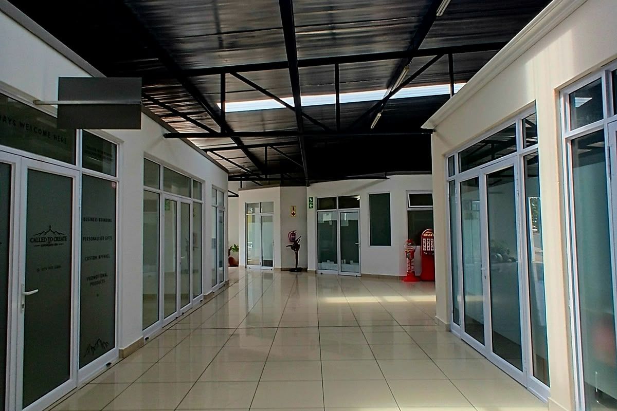 A bright, modern commercial hallway featuring tiled flooring, aluminum-framed glass office partitions, and an exposed steel roof structure with natural light panels.